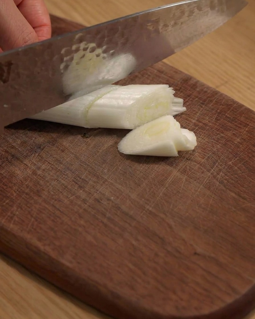 Ein Messer schneidet eine große Frühlingszwiebel auf einem Holzbrett in Stücke.