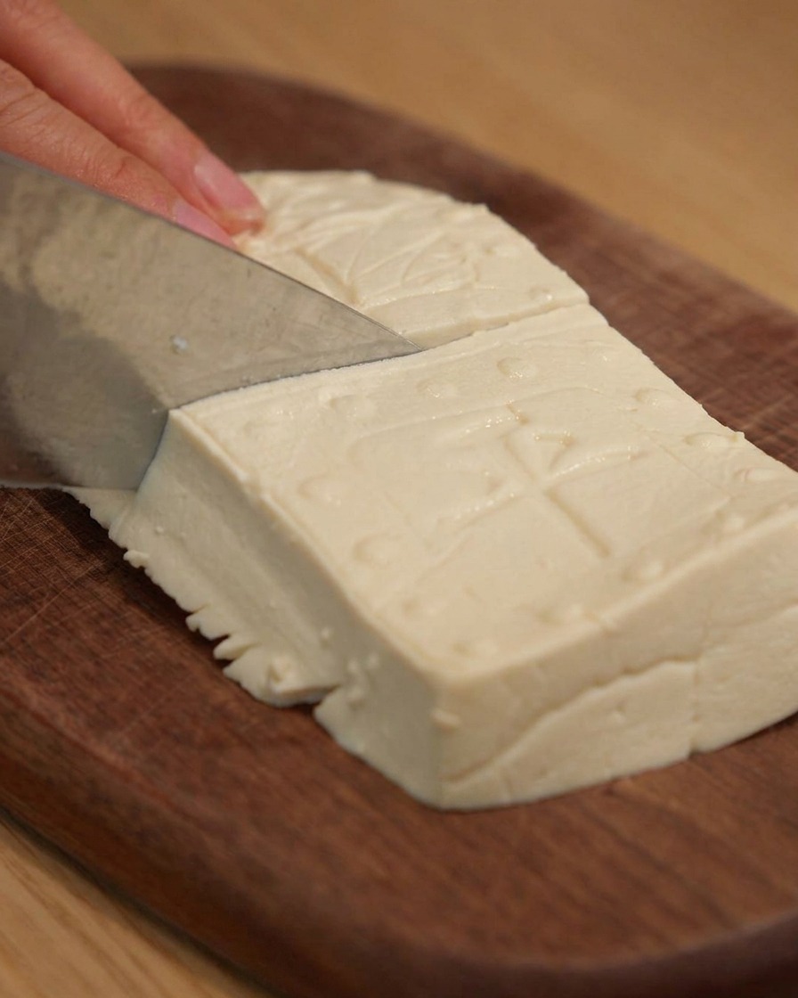 Ein scharfes Messer schneidet festen Tofu auf einem Holzschneidebrett in saubere rechteckige Blöcke.