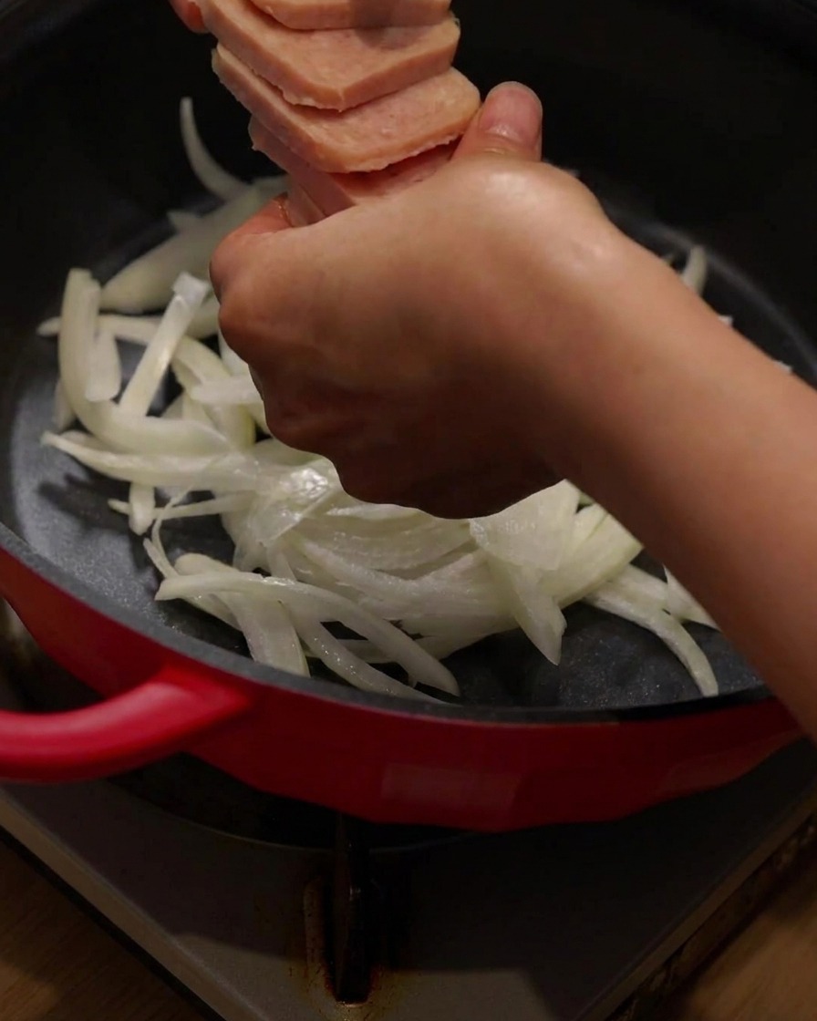 Geschnittenes Lunchmeat und Speck werden im roten Kochtopf auf den Zwiebeln angeordnet.
