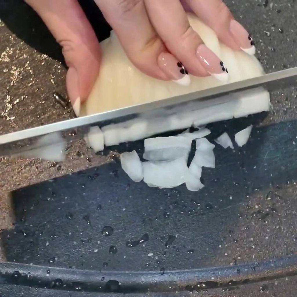 Eine Hand schneidet mit einem Messer eine weiße Zwiebel auf einem dunklen Schneidebrett, gehackter Knoblauch liegt daneben.