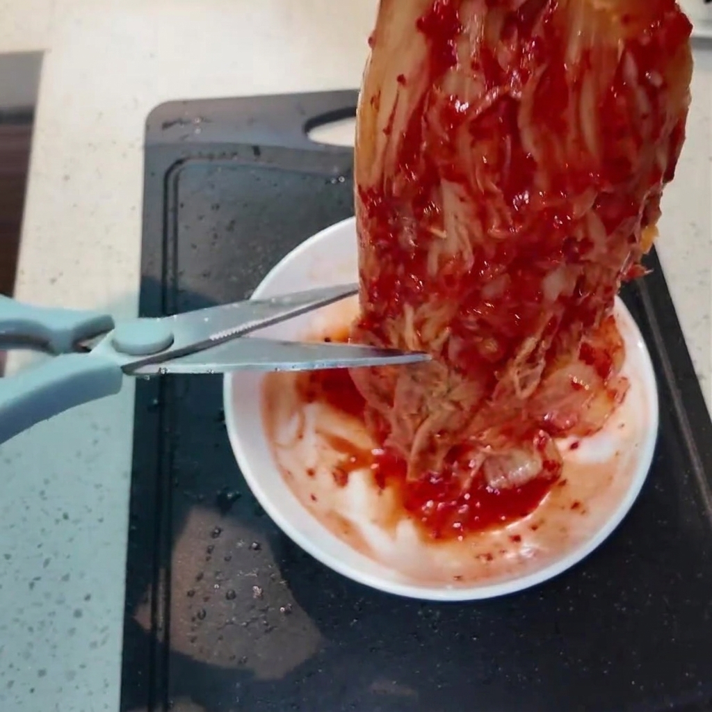 Ein Stück leuchtend roter Kimchi wird mit einer Küchenschere über einem kleinen weißen Teller in kleinere Stücke geschnitten.