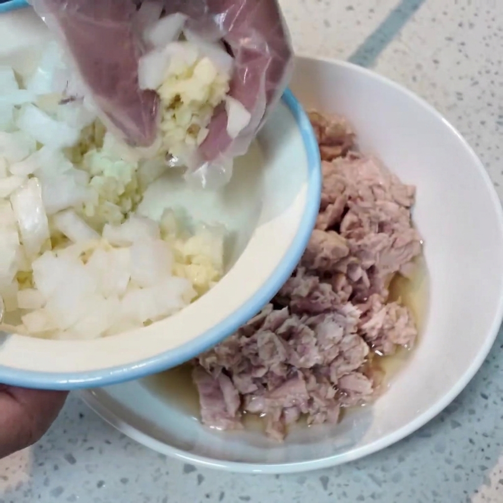 Eine handgeschützte Hand gibt gehackte Zwiebeln und Knoblauch aus einer kleinen Schüssel in eine größere Schüssel mit Thunfisch.