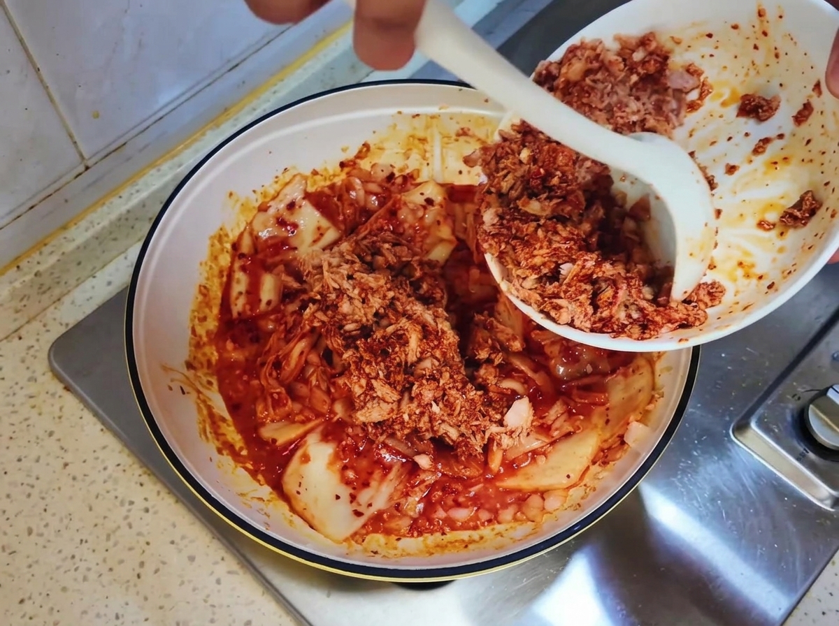 Die Thunfischmischung wird mit einem weißen Löffel aus einer Schüssel in die Pfanne zum Kimchi gegeben.