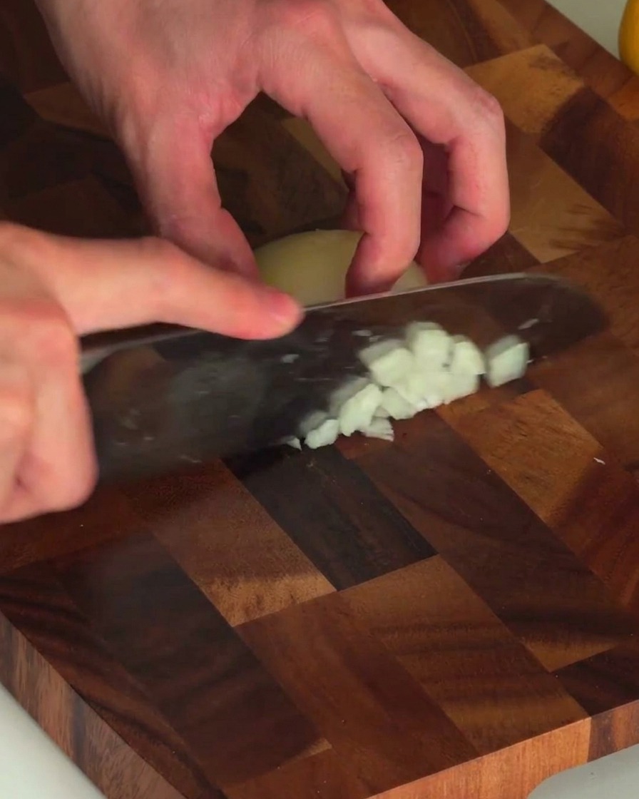 Hände halten ein Messer und hacken fein eine weiße Zwiebel auf einem Holzbrett.