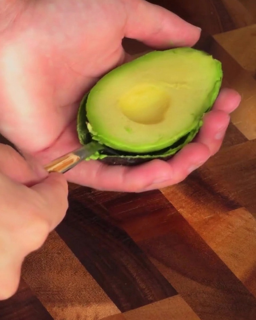 Eine Person löffelt mit einem Löffel das grüne Fruchtfleisch aus einer Avocadohälfte.
