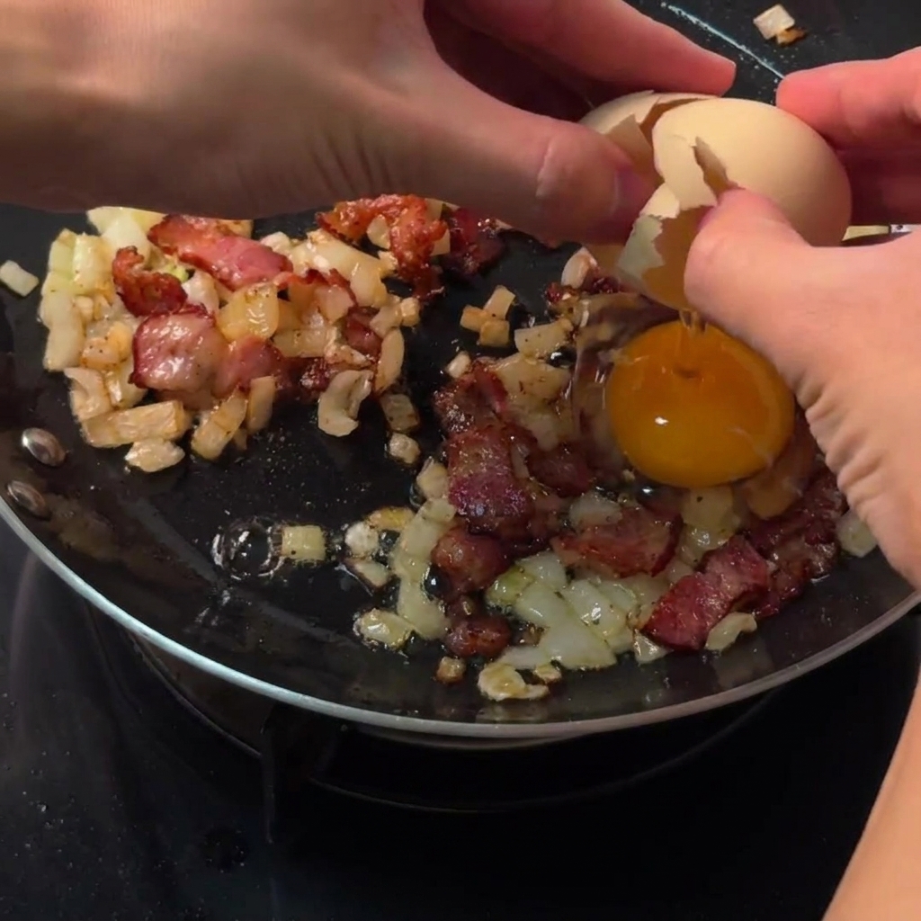 Hände schlagen ein rohes Ei direkt in eine freigemachte kreisförmige Lücke zwischen Speck und Zwiebeln in einer Pfanne.