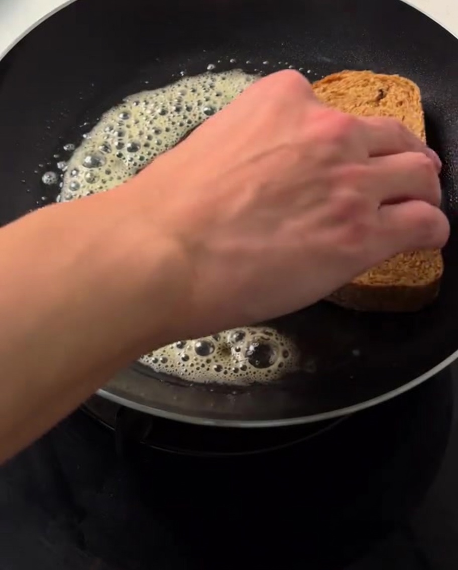 Eine Hand legt eine Scheibe Braunbrot auf blubbernde, geschmolzene Butter in einer heißen Pfanne.