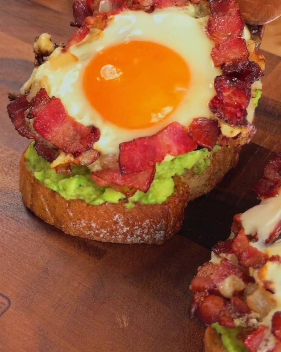Ein gebratener Speck- und Eierring wird mit einem Holzspatel auf das Avocadotoast geschichtet.
