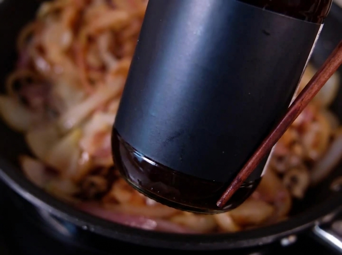 Eine dunkle Flasche, aus der Sauce über dünn geschnittene, gebräunte Zwiebeln in einer Pfanne gegossen wird.