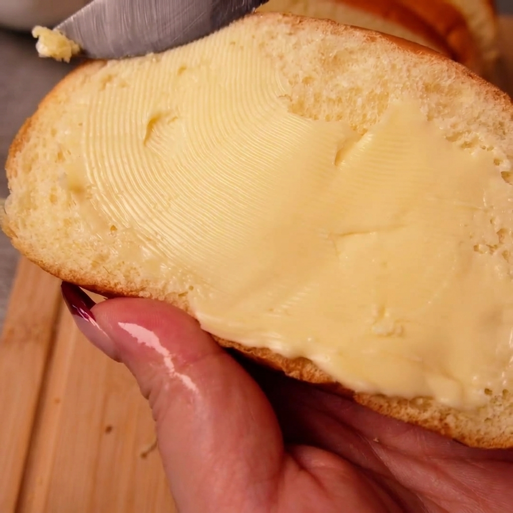 Eine Hand, die eine Scheibe Weißbrot hält, während mit einem Messer Butter gleichmäßig auf der Oberfläche verteilt wird.
