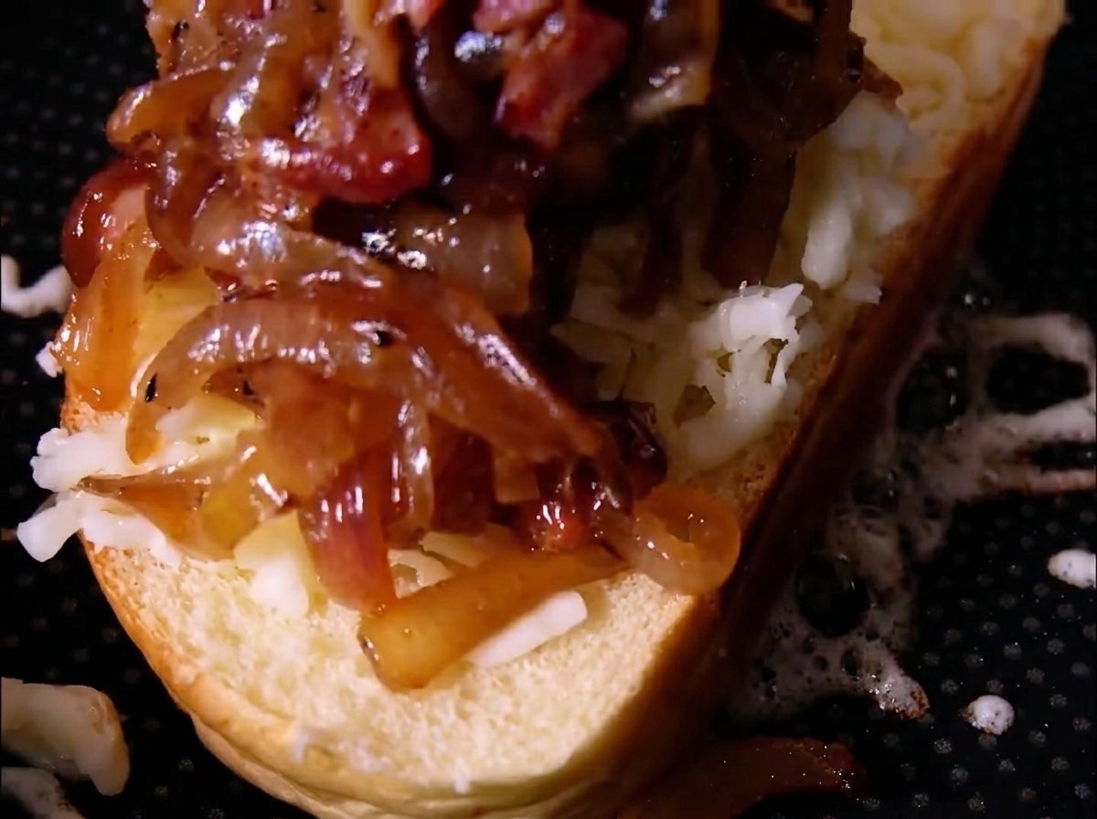 Ein großzügiger Haufen karamellisierter Zwiebeln und Speckstücke liegt auf geriebenem Käse auf einer Brotscheibe.