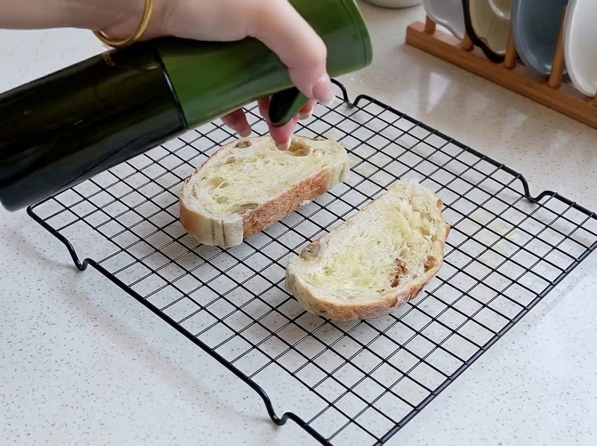 Eine Hand sprüht eine feine Schicht Öl auf zwei Scheiben rustikales Brot, die auf einem Kuchengitter liegen.
