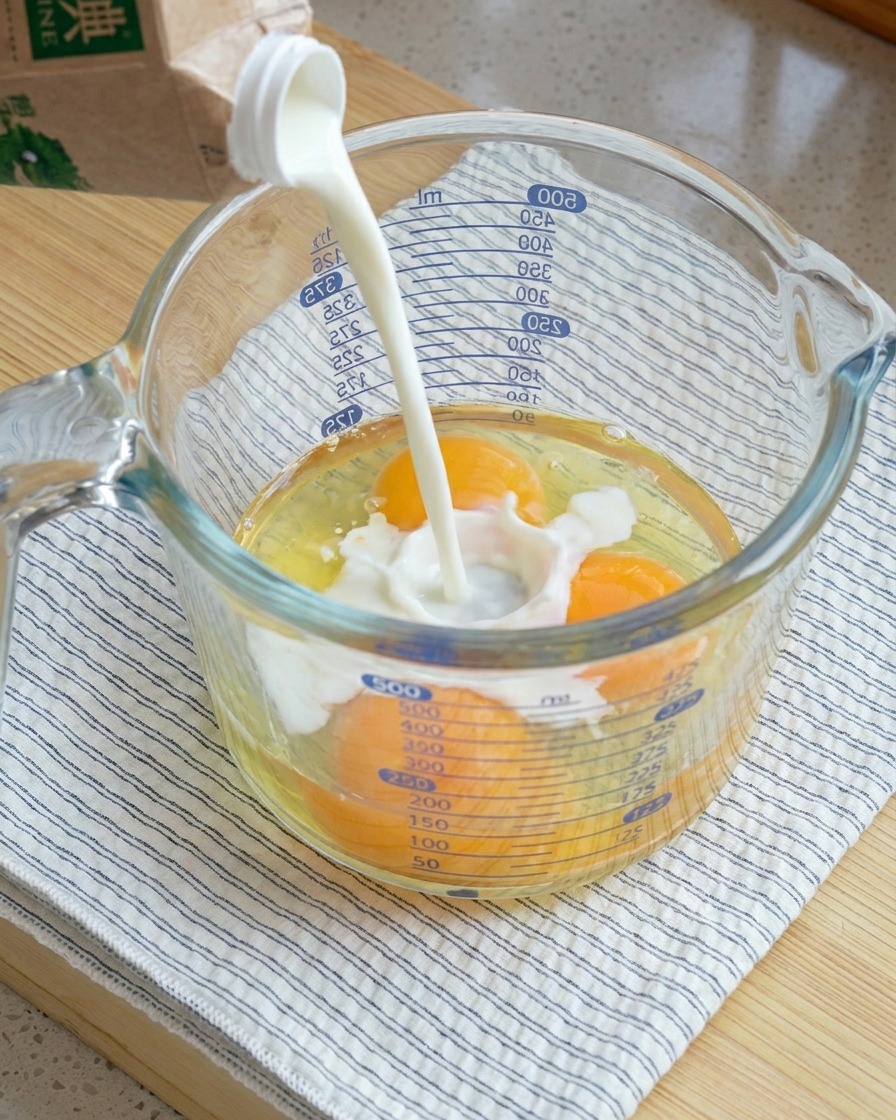 Milch wird in einen großen Glasmessbecher mit drei aufgeschlagenen rohen Eiern gegossen.