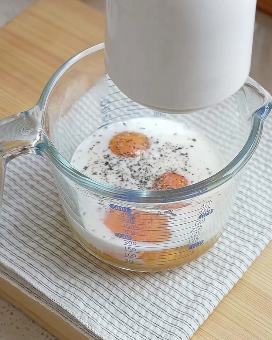 Ein Milchaufschäumer wird über einen Glasmessbecher gehalten, der rohe Eier, Milch und frisch gemahlenen schwarzen Pfeffer enthält.