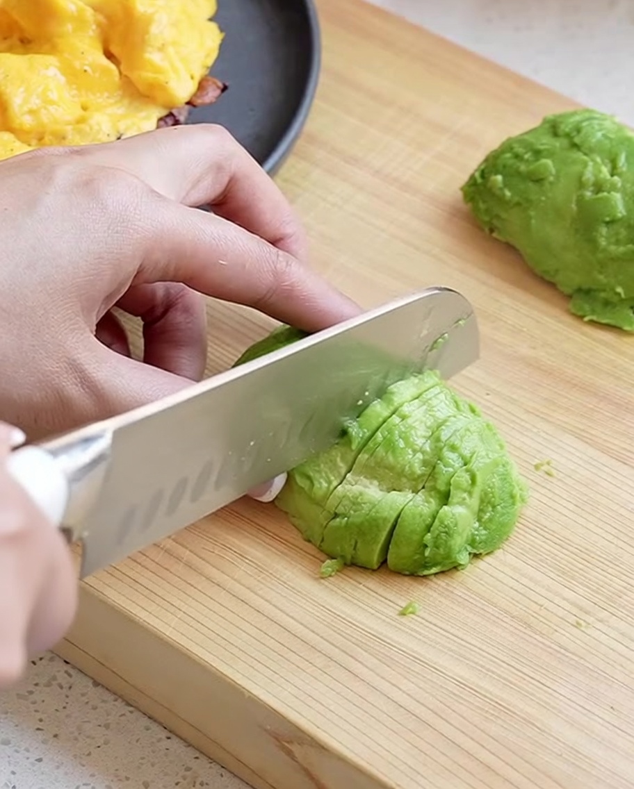 Eine Hand schneidet mit einem Chefmesser eine geschälte Avocadofälfte in saubere, dünne Scheiben auf einem Holzbrett.