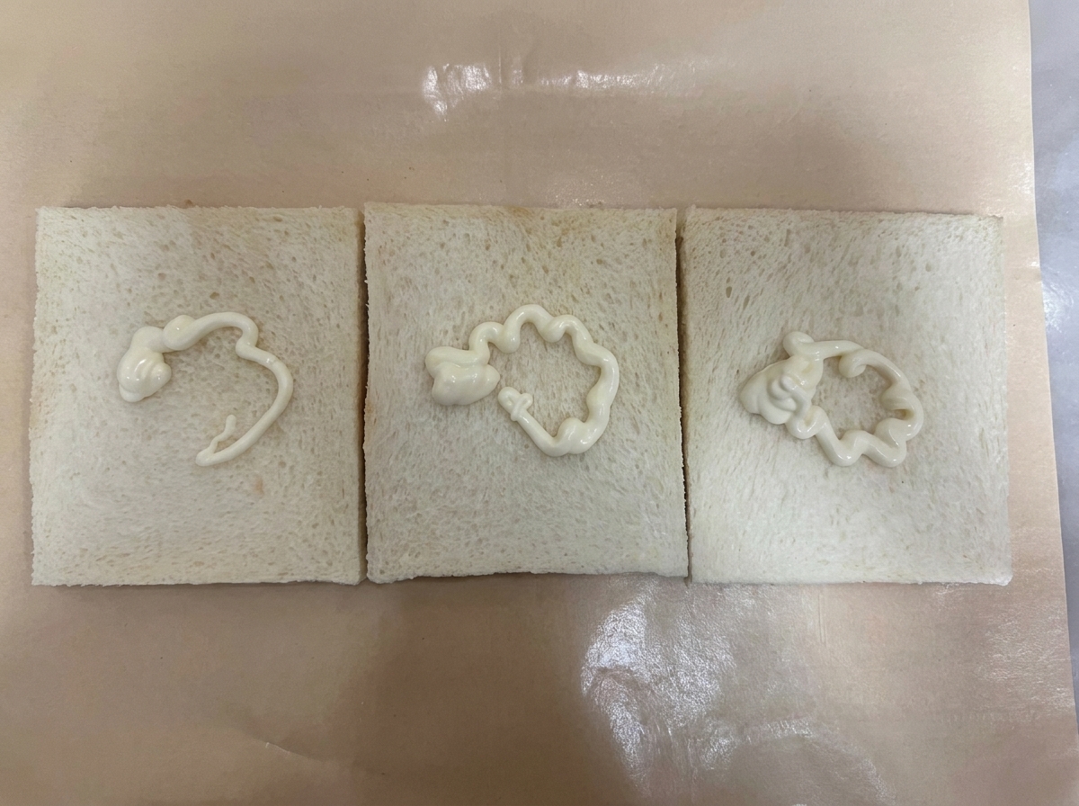 Drei Scheiben krustenloses Weißbrot nebeneinander auf braunem Backpapier, jeweils mit einem geschwungenen Streifen weißem Salatdressing versehen.