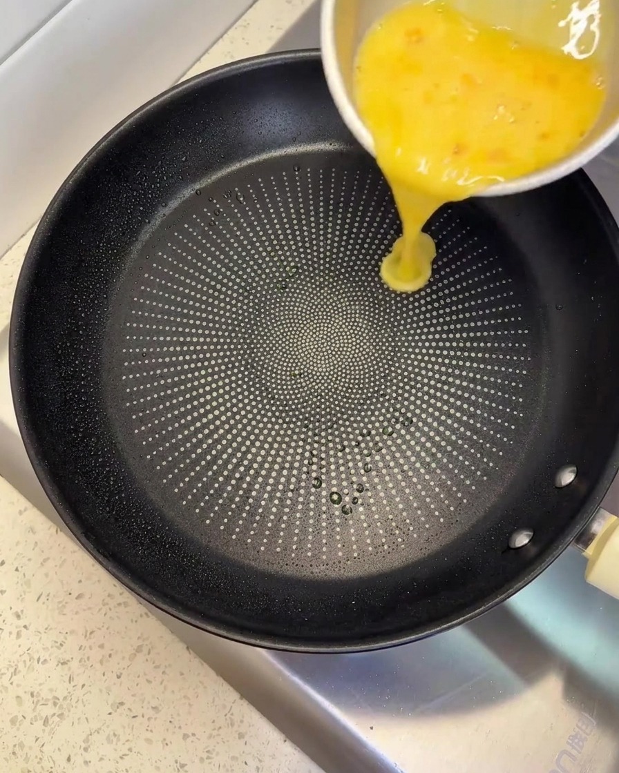 Das Eingießen einer gelben, geschlagenen Eimischung aus einer Metallschüssel in eine schwarze Antihaftpfanne.