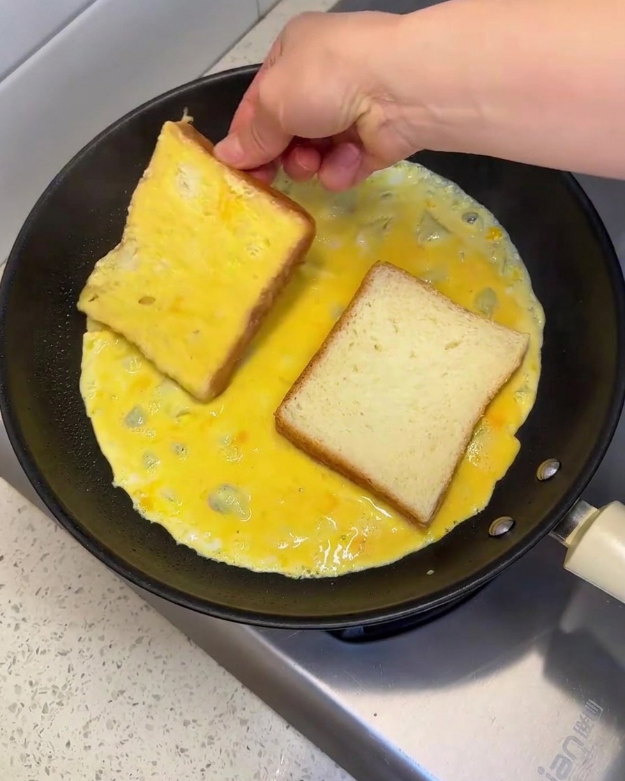 Eine Hand platziert und wendet eine Brotscheibe in einer heißen Pfanne mit rohem, verquirltem Ei, um beide Seiten zu bedecken.