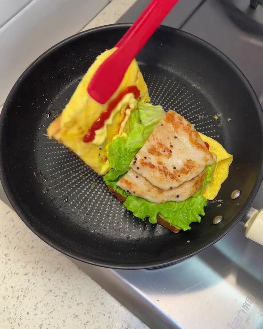 Ein Paar rote Zangen hebt und faltet eine Seite des Brots, um das Sandwich in der Pfanne zu schließen.