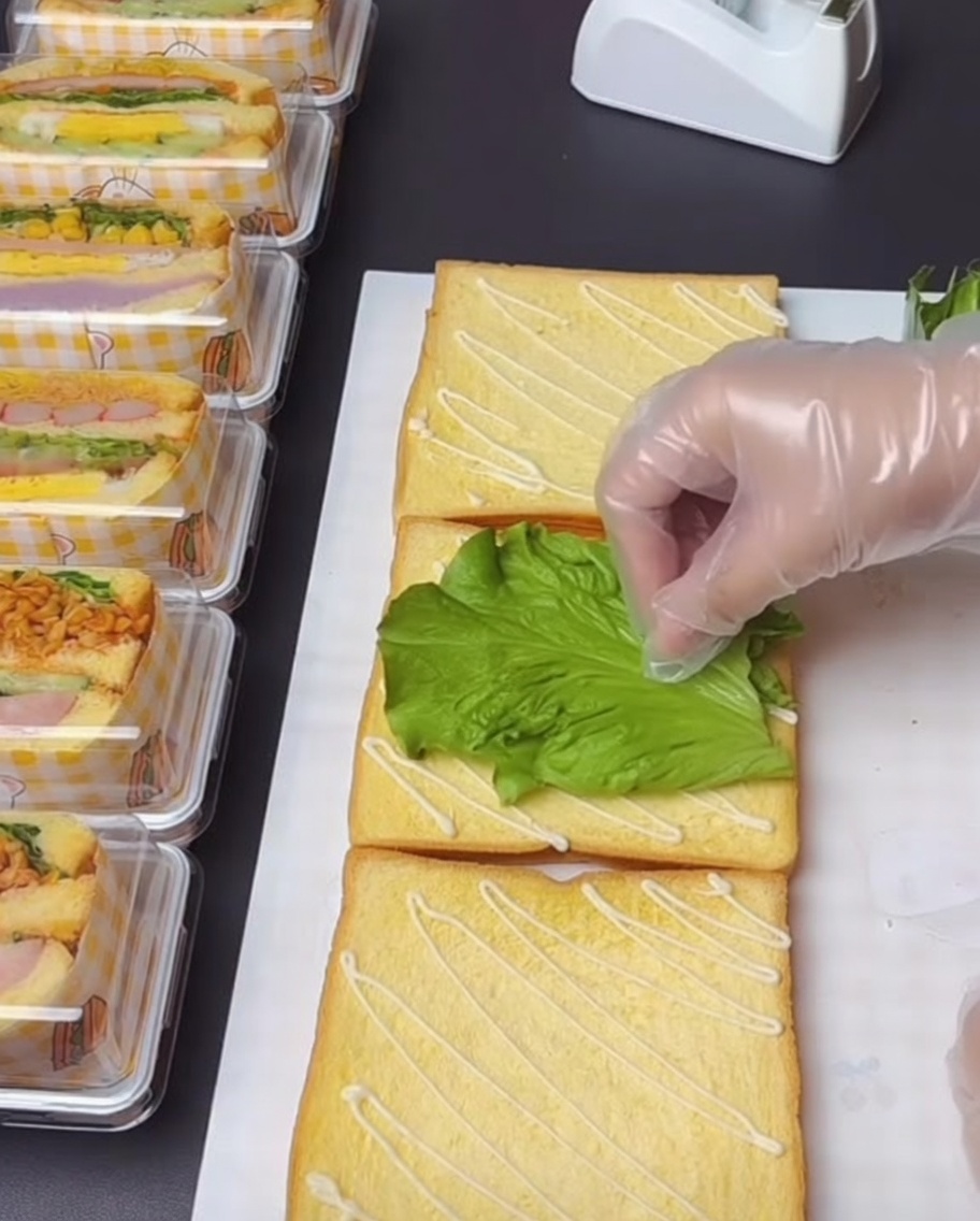 Eine behandschuhte Hand legt vorsichtig ein frisches, grünes Salatblatt auf eine mit Salatdressing beträufelte Toastscheibe.