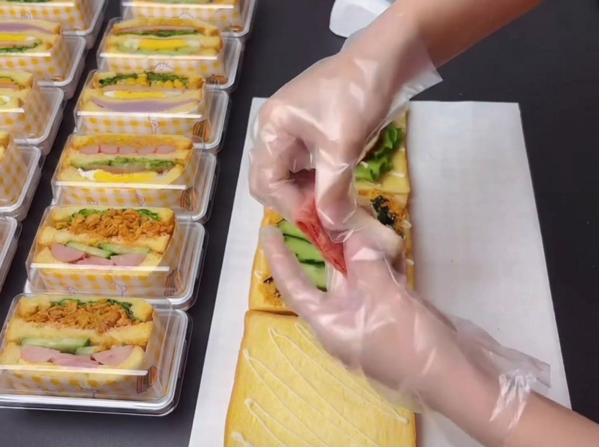 Behandschuhte Hände halten und arrangieren kleine Stücke gebratenen Specks, um sie auf das Sandwich zu legen.