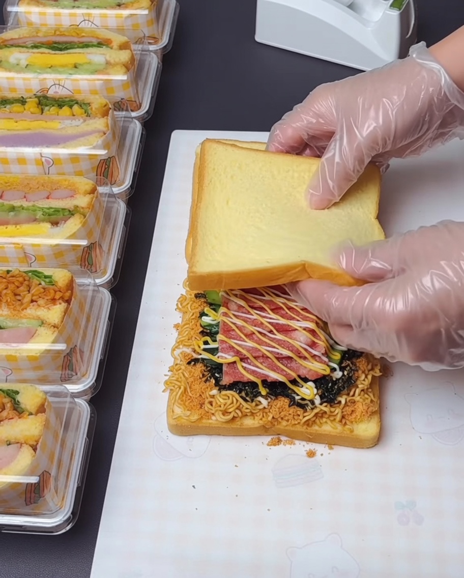 Ein Paar behandschuhte Hände hebt die oberste Toastscheibe an und stapelt sie auf die darunter liegenden, belegten Schichten.