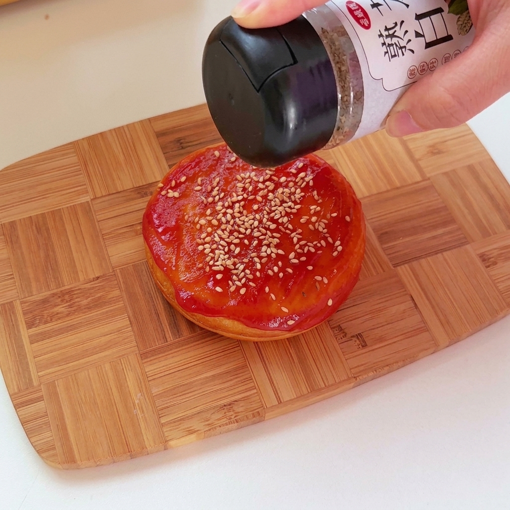 Eine Nahaufnahme von Ketchup, der auf die Oberseite eines herzhaften, runden Brötchens mit Sesam aufgetragen wird.