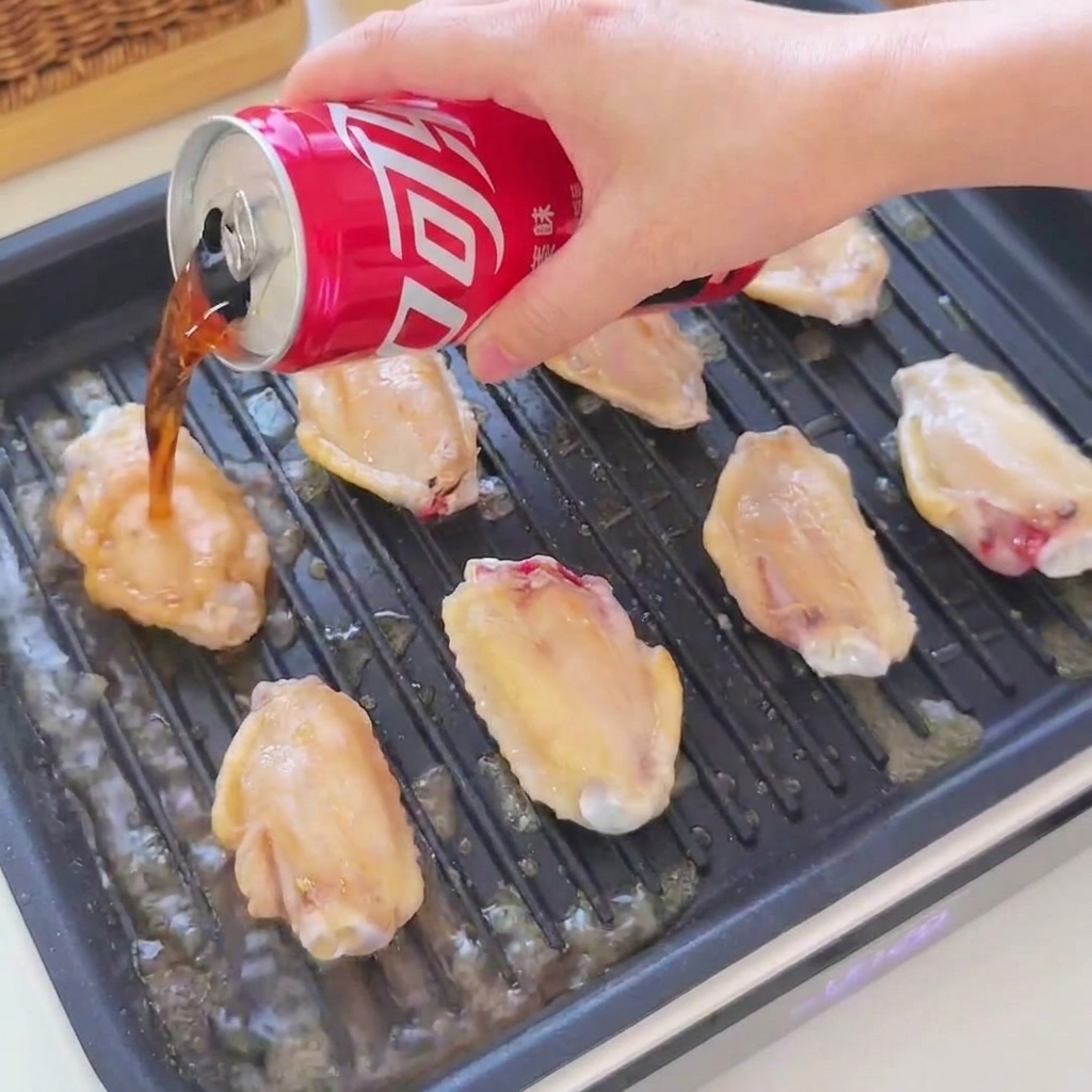 Cola und Sojasauce werden aus einer roten Dose über goldene Hähnchenflügel in einer schwarzen Grillpfanne gegossen.