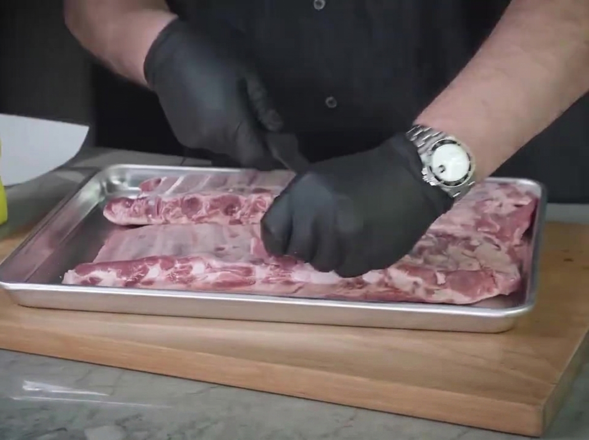 Eine Nahaufnahme von behandsholten Händen, die überschüssiges Fett und zähe Silberhaut auf einem Stahlblech von rohen St. Louis Ribs entfernen.