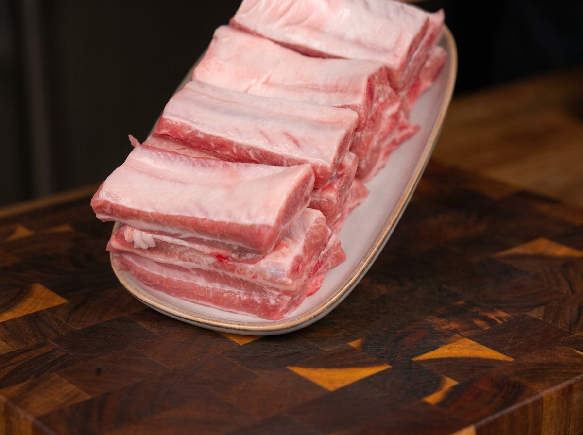 Frische, rohe Schweinerippchen säuberlich auf einem ovalen weissen Teller auf einem Holzschneidebrett angeordnet.