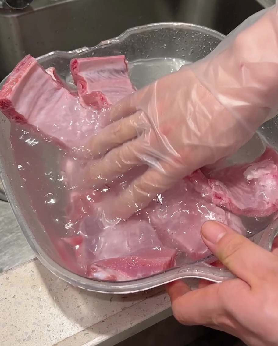 Mit Handschuhen bekleidete Hände schrubben und waschen rohe Schweinerippchen in einem wassergefüllten Behälter.