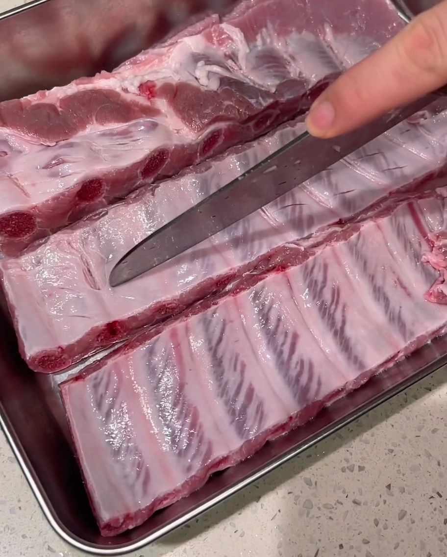Ein scharfes Messer schneidet flache Schnitte in die Silberhaut von rohen Schweinerippchen auf einem Metalltablett.