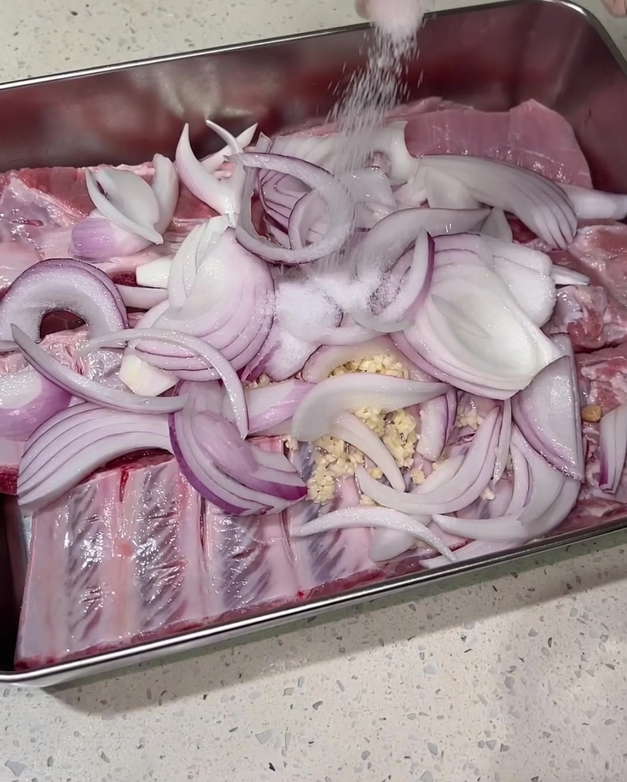 Salz wird großzügig mit der Hand über die gewürzten Schweinerippchen und Zwiebeln gestreut.