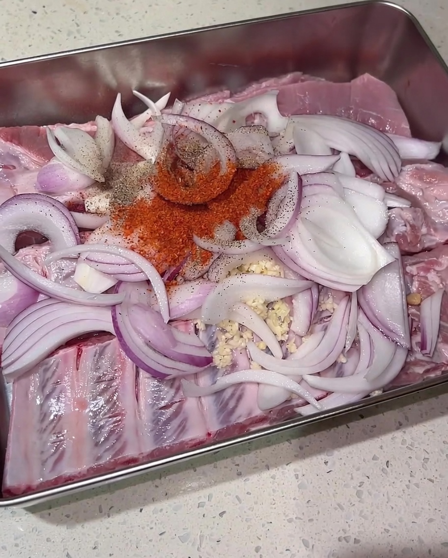 Ein Häufchen rotes Chilipulver, schwarzer Pfeffer und brauner Zucker liegen auf rohen Schweinerippchen und roten Zwiebeln.
