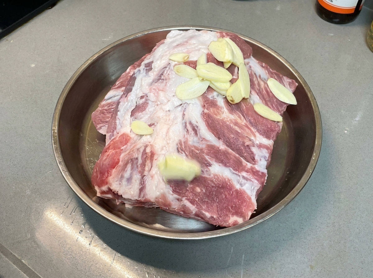Knoblauchscheiben liegen gleichmäßig auf rohen Schweinerippchen in einer Metallschale.