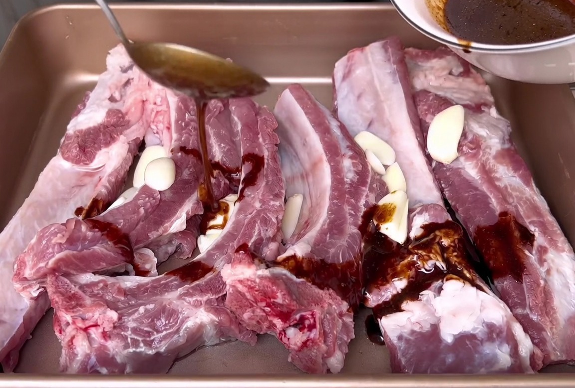 Dunkle Soja-Marinade wird aus einer weißen Schüssel über rohe Schweinerippchen und Knoblauchscheiben in einer Backform gegossen.