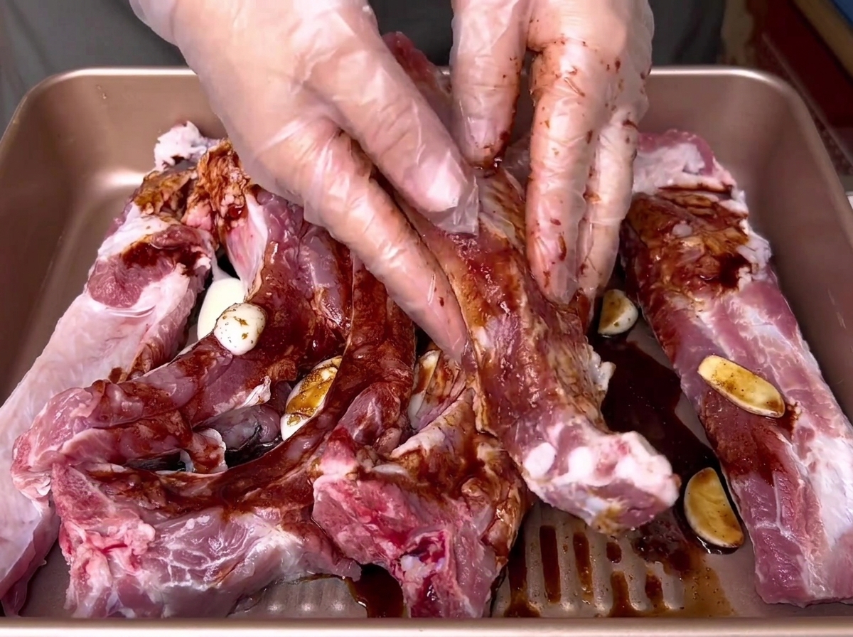 Mit Handschuhen bekleidete Hände reiben eine dunkle flüssige Marinade in einer goldenen Form gründlich in rohe Schweinerippchen ein.