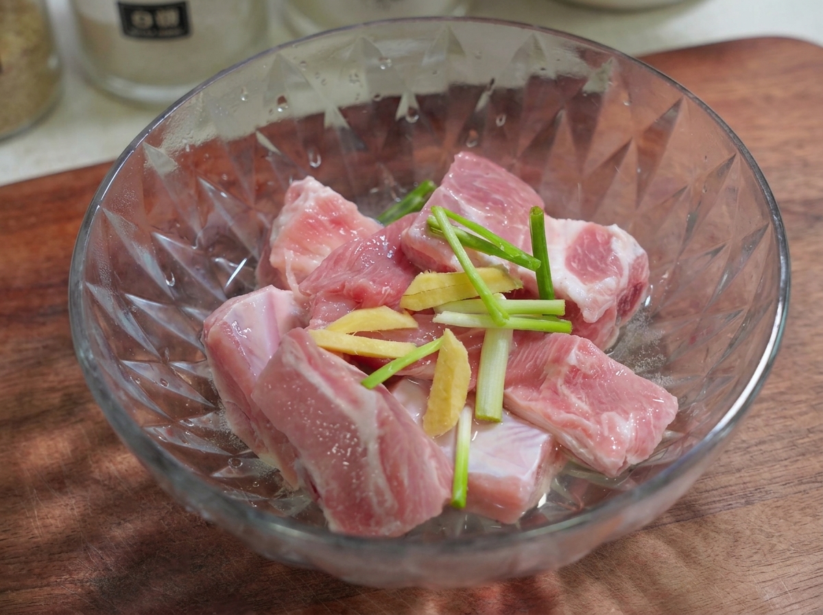 Geschnittene Ingwer- und Frühlingszwiebelstücke liegen oben auf rohen Schweinerippchen in einer Glasschüssel.