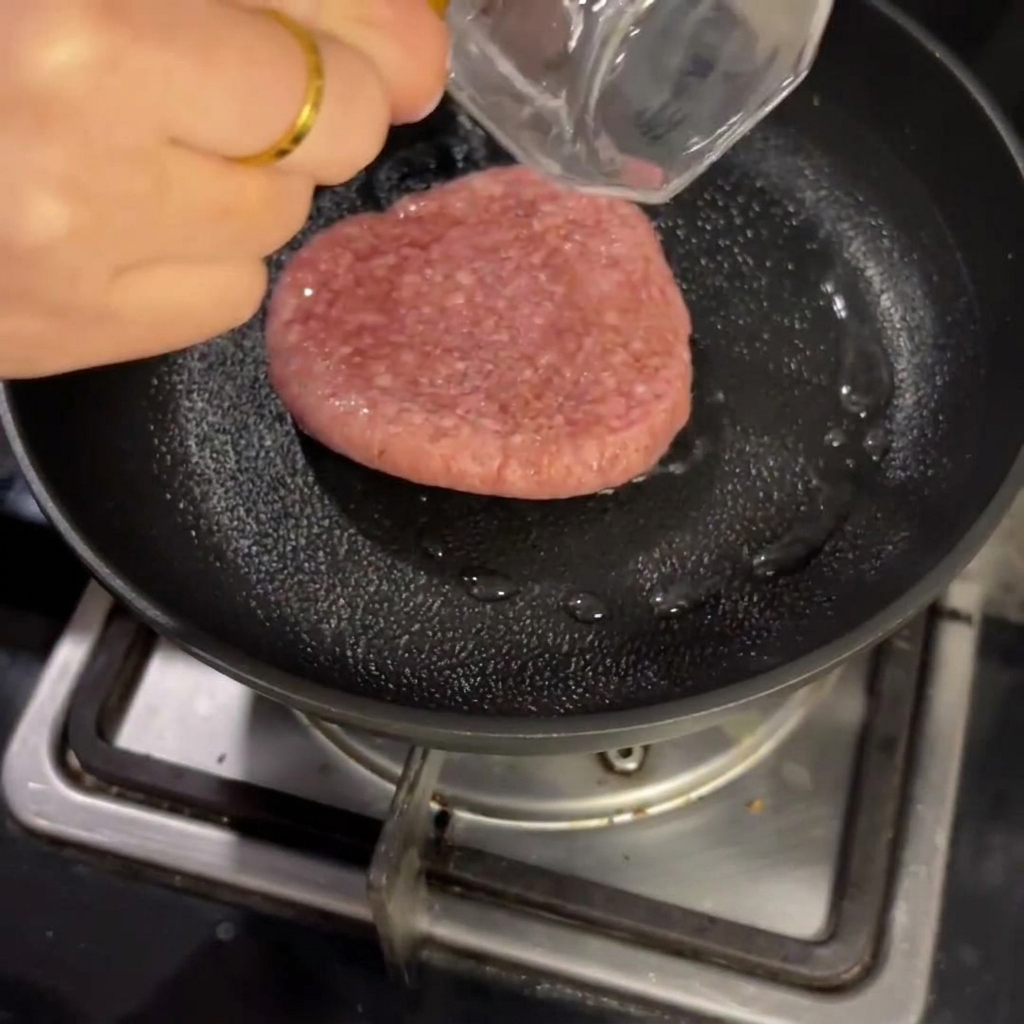 Ein rohes Rinderpatty brät in einer Pfanne mit etwas Flüssigkeit, abgedeckt mit einem Deckel.