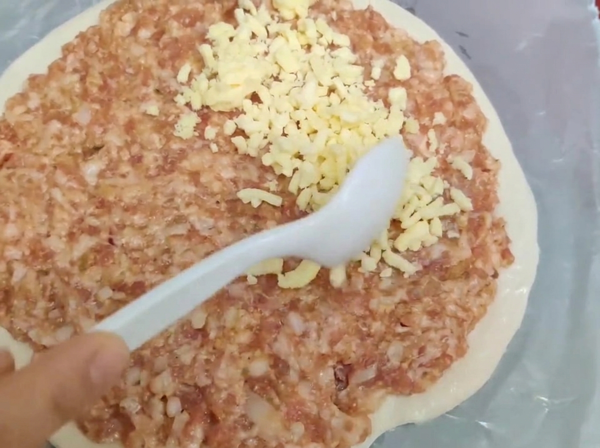 Ein weißer Löffel verteilt rohes Hackfleisch und geriebenen Käse gleichmäßig auf einem runden Teigboden.