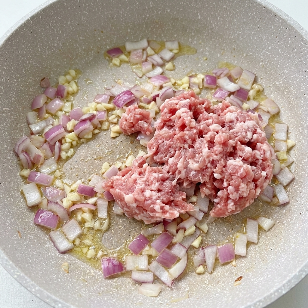 Rohes Schweinehackfleisch in einer Pfanne neben garen Zwiebeln und gehacktem Knoblauch.