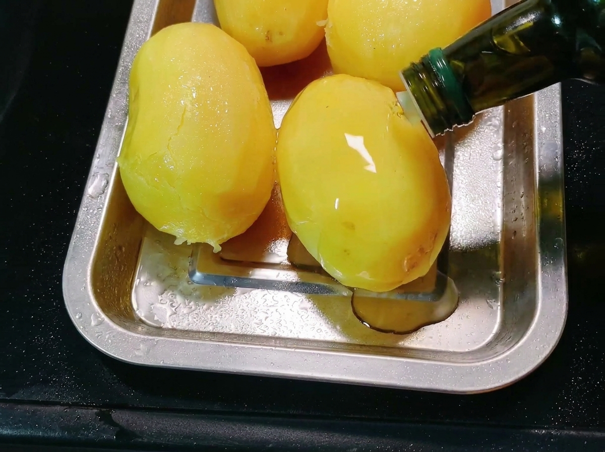 Speiseöl wird aus einer Flasche auf geschälte Kartoffeln in einem Backblech aus Metall gegossen.