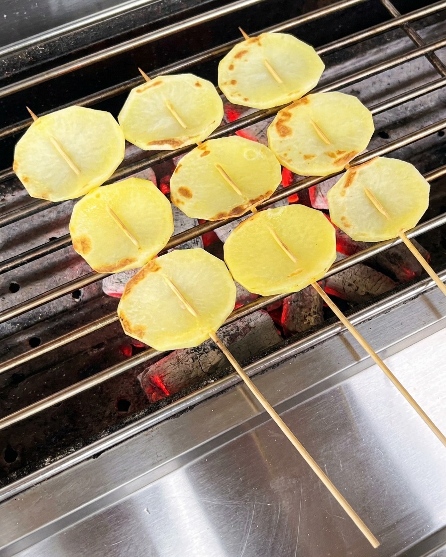 Teilweise gegarte Kartoffelscheiben auf dem Grill, die mit einem Pinsel eingeölt werden.