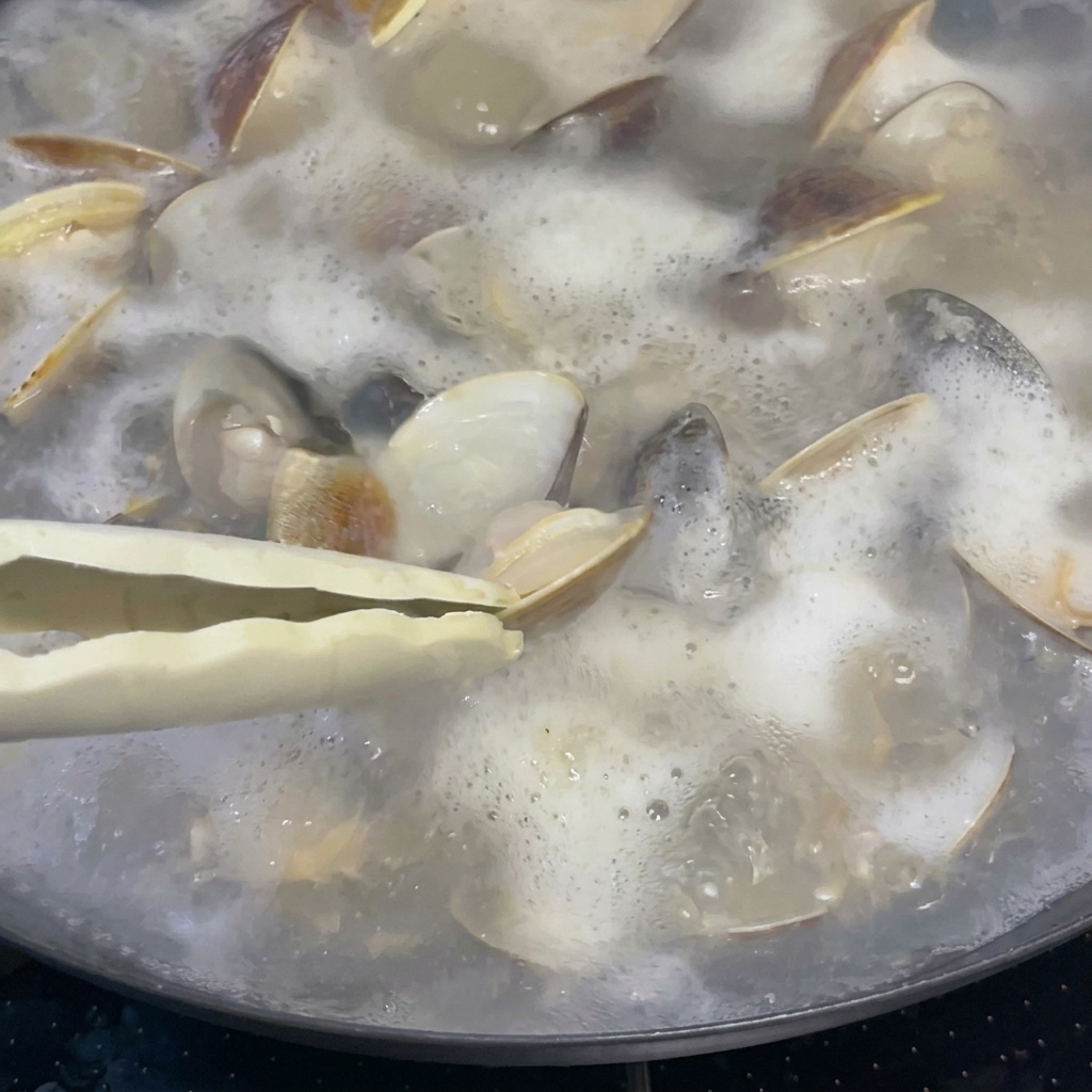 Ein Esslöffel Reiswein wird in einen Topf mit sprudelnden Muscheln und Brühe gegossen.