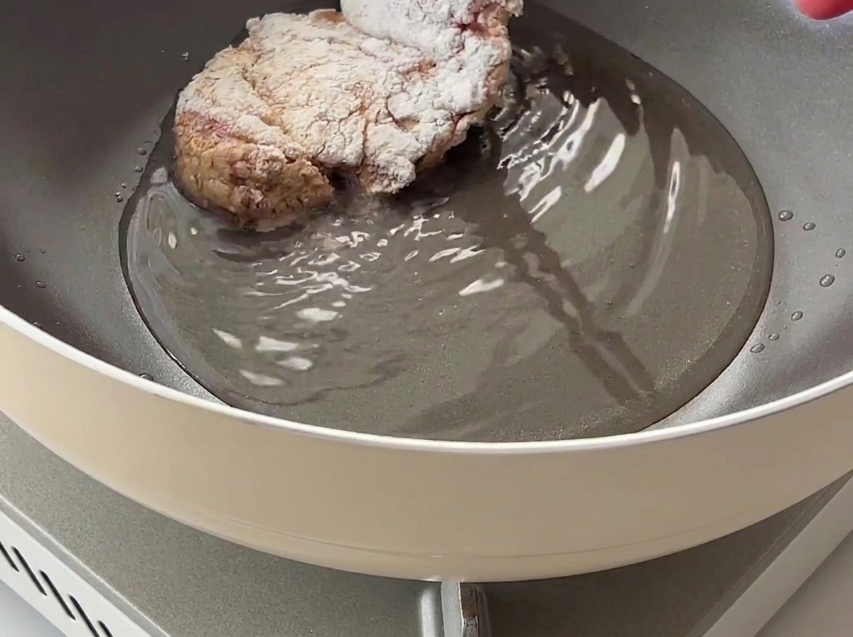 Ein mit Maisstärke ummantelter Hähnchenschenkel wird in eine Pfanne mit heißem Speiseöl gelegt.