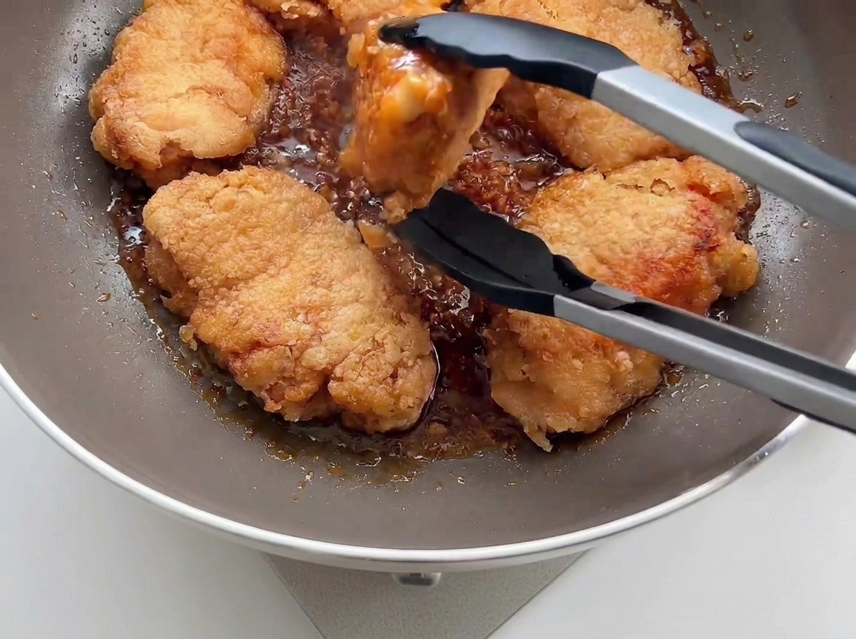 Knusprig frittiertes Hähnchen wird mit einer schwarzen Küchenzange in eine Pfanne mit Knoblauchsauce gegeben.