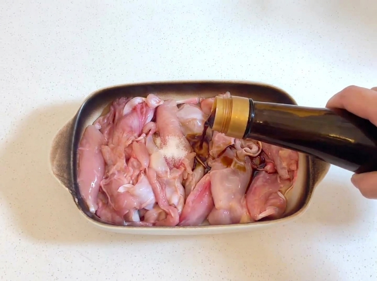 Dunkle Sojasauce wird aus einer Glasflasche auf rohe Hähnchenstücke gegossen, wobei eine kleine Prise Salz auf dem Hähnchen sichtbar ist.
