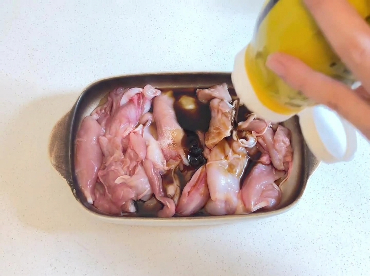 Dickflüssige, dunkle Austernsauce wird aus einer gelben Flasche auf rohe Hähnchenstücke gedrückt, die in einer Sojasaucenmarinade liegen.