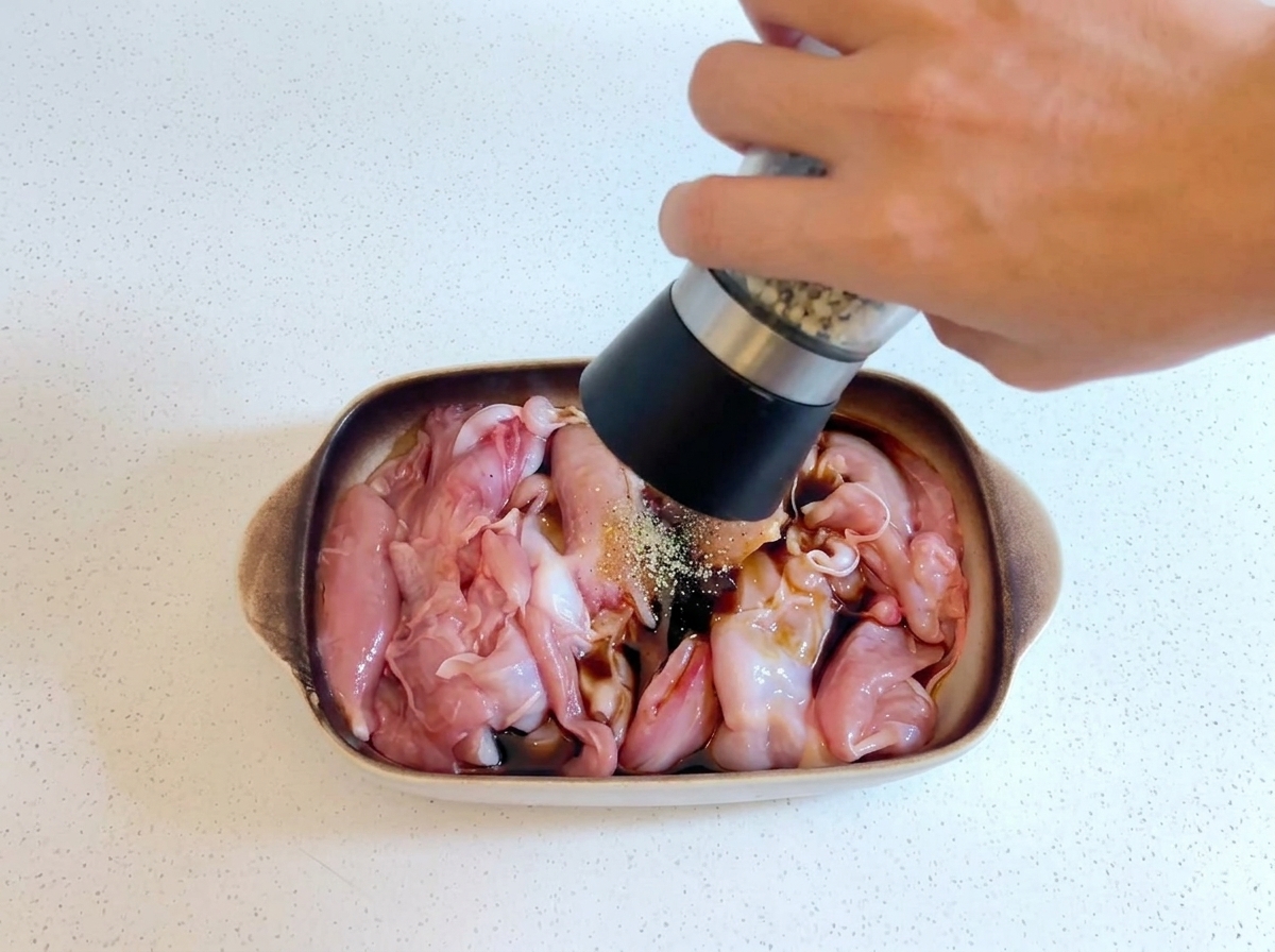 Eine Hand hält eine Pfeffermühle und mahlt frischen Pfeffer über rohes Hähnchen in einer Schüssel.