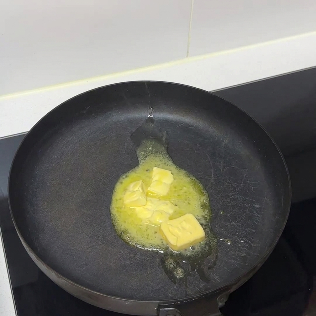 Butterstücke schmelzen in einer dunklen Bratpfanne auf einem Herd zu Flüssigkeit.